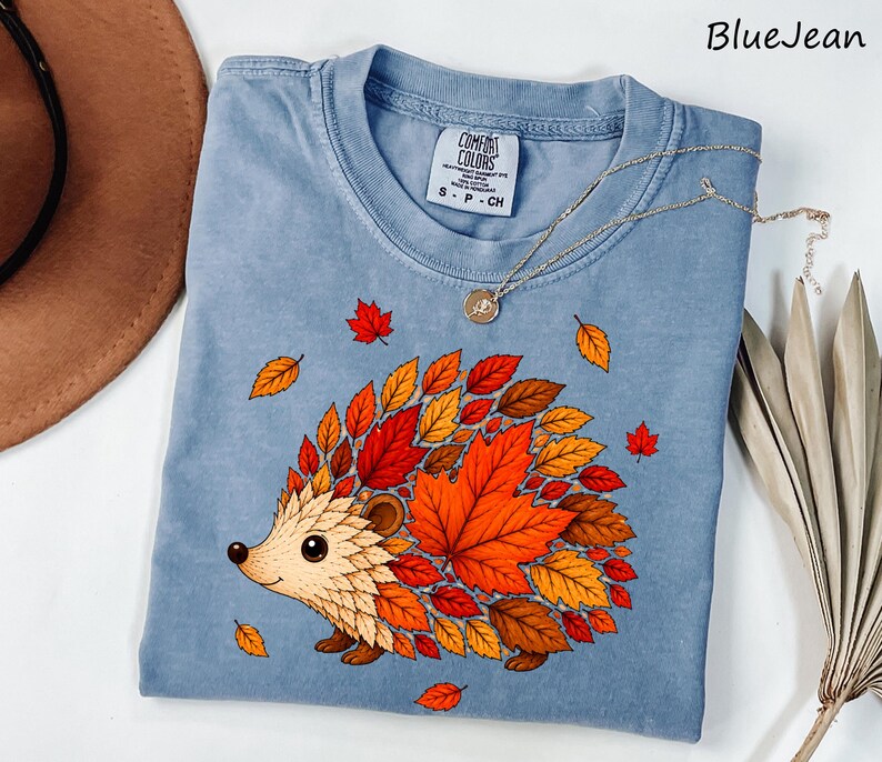 May include: A blue jean colored t-shirt featuring a graphic of a hedgehog whose back is covered in vibrant autumn leaves in shades of red, orange, and brown. The shirt has a crew neckline and a Comfort Colors brand label on the inside collar.