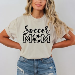 May include: A beige t-shirt with the text "Soccer Mom" in black lettering and a black and white soccer ball graphic.