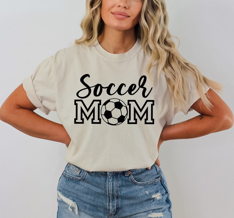 May include: A beige t-shirt with the text "Soccer Mom" in black lettering and a black and white soccer ball graphic.