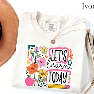 May include: Ivory-colored t-shirt with a floral design and the text "LET'S learn TODAY". The design includes an apple, pencil, and various flowers in pink, orange, yellow, and green. A gold necklace is draped across the shirt.