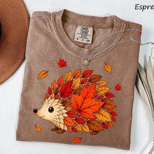 May include: An espresso-colored t-shirt featuring a graphic of a hedgehog whose back is covered in vibrant red, orange, and yellow autumn leaves. The shirt has a crew neckline and a Comfort Colors brand label on the inside collar area.