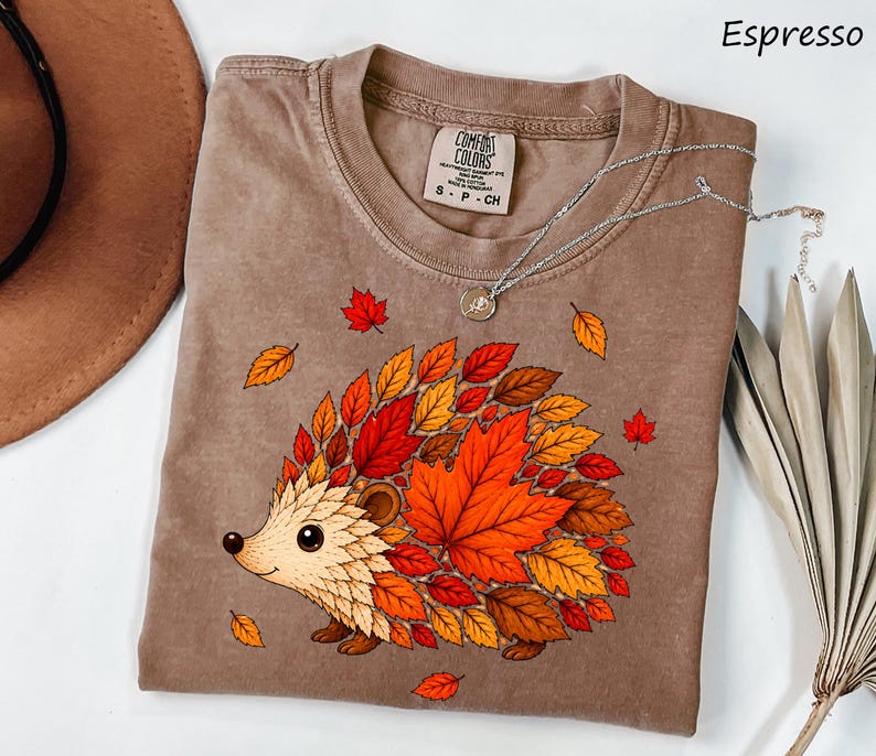 May include: An espresso-colored t-shirt featuring a graphic of a hedgehog whose back is covered in vibrant red, orange, and yellow autumn leaves. The shirt has a crew neckline and a Comfort Colors brand label on the inside collar area.