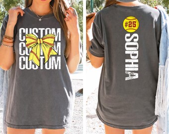 May include: Gray t-shirt with a softball bow graphic and the word "CUSTOM" repeated three times. The back of the shirt features a softball graphic with the number "25" and the name "SOPHIA" printed vertically.