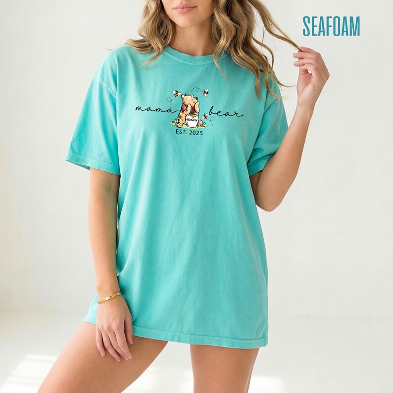 May include: Seafoam green t-shirt with a graphic of Winnie the Pooh holding a honey pot, surrounded by bees. The text "mama bear" is above the graphic, with "EST. 2025" below. The shirt is a loose fit.