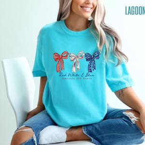May include: A bright turquoise t-shirt featuring a graphic of three bows in red gingham, white with blue stripes, and blue with white stars. Below the bows, the text reads Red White & Blue America 250 Years. The shirt is paired with blue jeans.