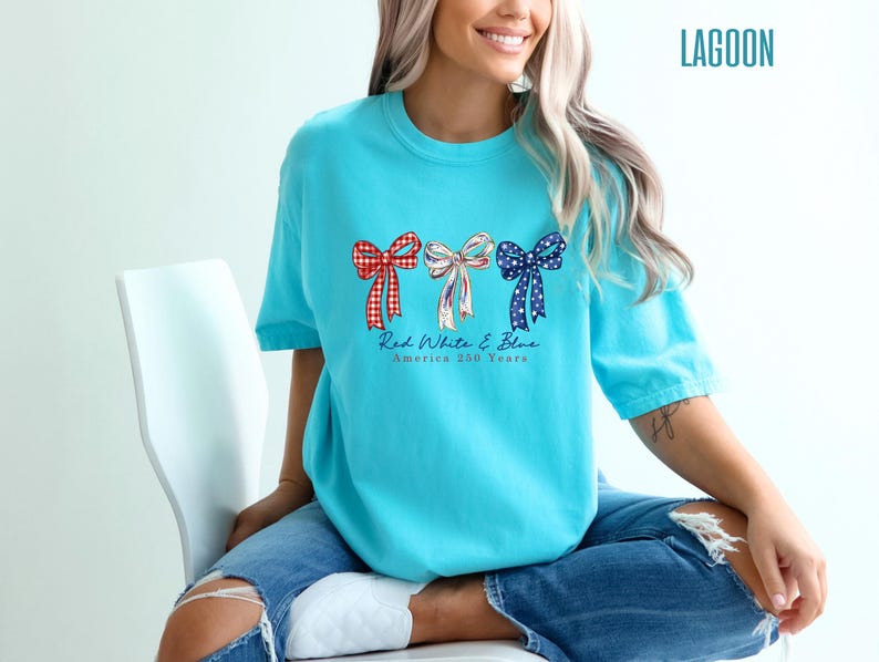 May include: A bright turquoise t-shirt featuring a graphic of three bows in red gingham, white with blue stripes, and blue with white stars. Below the bows, the text reads Red White & Blue America 250 Years. The shirt is paired with blue jeans.