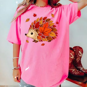 May include: A bright pink short-sleeved t-shirt featuring a graphic of a hedgehog with a back made of colorful autumn leaves in shades of red, orange, and yellow. The text CRUNCHBERRY is printed in black in the upper left corner of the image.