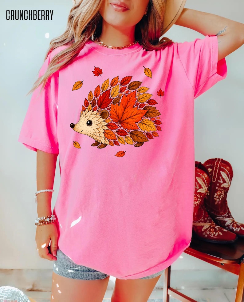 May include: A bright pink short-sleeved t-shirt featuring a graphic of a hedgehog with a back made of colorful autumn leaves in shades of red, orange, and yellow. The text CRUNCHBERRY is printed in black in the upper left corner of the image.
