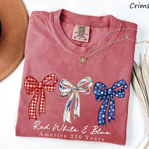 May include: A crimson short-sleeve t-shirt featuring three bows in red gingham, white with blue accents, and blue with white stars. Below the bows, the text reads Red White & Blue America 250 Years. The shirt has a Comfort Colors brand label at the neck.