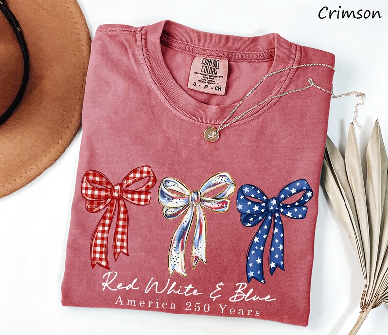May include: A crimson short-sleeve t-shirt featuring three bows in red gingham, white with blue accents, and blue with white stars. Below the bows, the text reads Red White & Blue America 250 Years. The shirt has a Comfort Colors brand label at the neck.