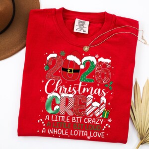 以下が含まれることがあります： 2026 Christmas Crew, A Little Bit Crazy, A Little Bit Loud, A Whole Lotta Loveというテキストがプリントされた赤いクルーネックTシャツ。デザインには、お祝いの模様、サンタの帽子、数字の0にあしらわれたベルトが含まれています。