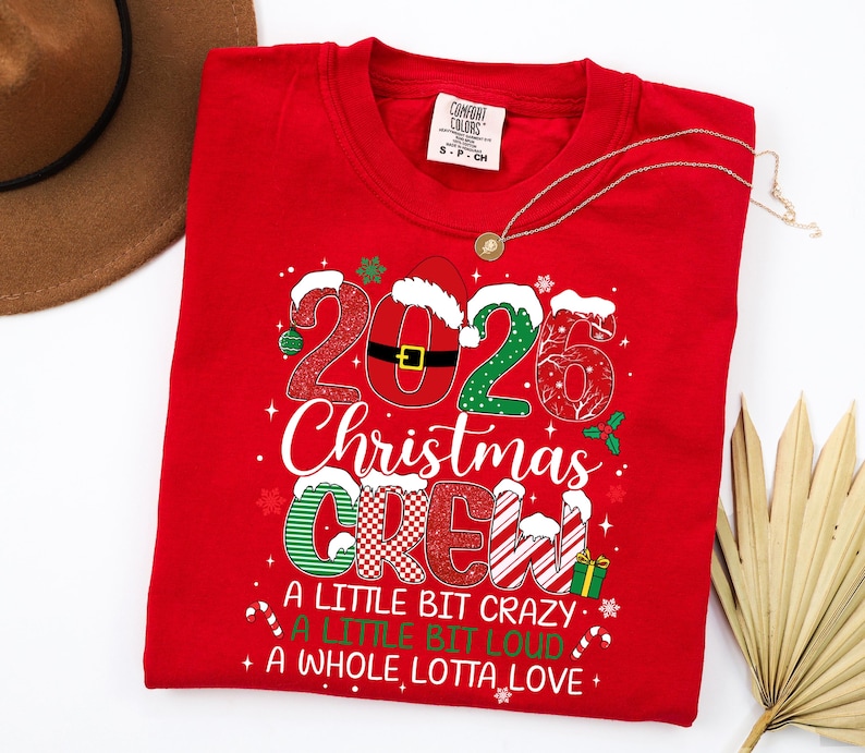以下が含まれることがあります： 2026 Christmas Crew, A Little Bit Crazy, A Little Bit Loud, A Whole Lotta Loveというテキストがプリントされた赤いクルーネックTシャツ。デザインには、お祝いの模様、サンタの帽子、数字の0にあしらわれたベルトが含まれています。