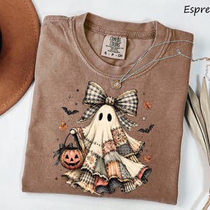 May include: An espresso-colored t-shirt featuring a graphic of a ghost wearing a large plaid bow and holding a jack-o'-lantern. The ghost is depicted with a patchwork-style body, surrounded by small bats, stars, and autumn leaves.