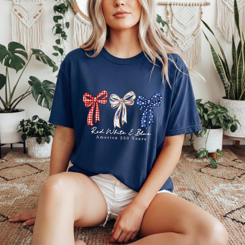 May include: A navy blue t-shirt featuring a graphic of three bows in red gingham, white with blue and red stripes, and blue with white stars. Below the bows, the text reads Red White & Blue America 250 Years in a white script font.