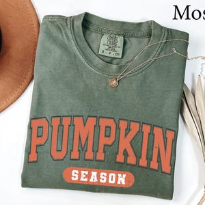 May include: A moss green t-shirt with the words "PUMPKIN SEASON" in orange block letters. The shirt has a crew neck and short sleeves. A gold necklace with a pendant is draped across the shirt. A brown hat and dried plant are in the background.