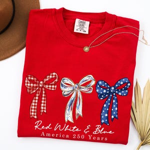 May include: A red crewneck t-shirt featuring three illustrated bows in red gingham, white with blue accents, and blue with white stars. Below the bows, the text reads Red White & Blue America 250 Years. The shirt has a Comfort Colors brand label.