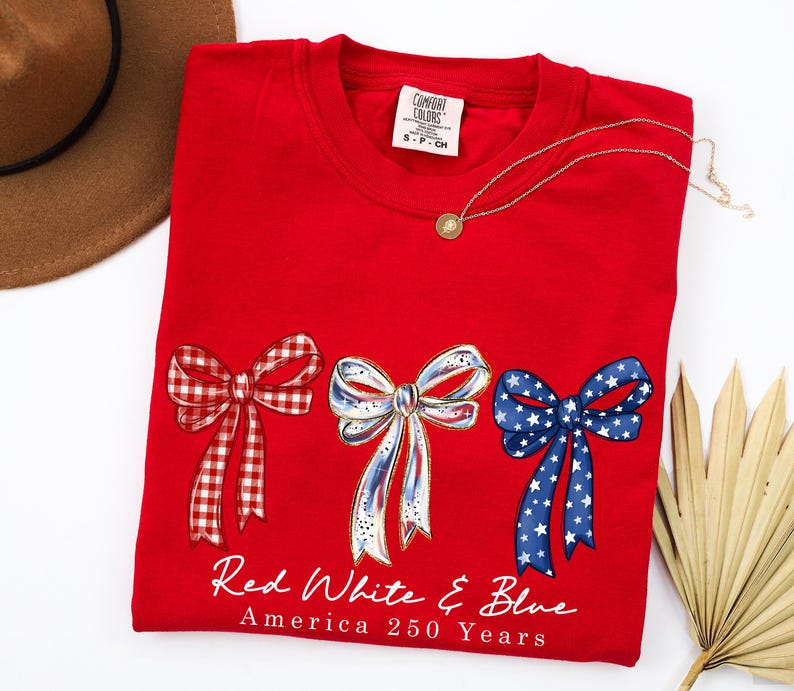May include: A red crewneck t-shirt featuring three illustrated bows in red gingham, white with blue accents, and blue with white stars. Below the bows, the text reads Red White & Blue America 250 Years. The shirt has a Comfort Colors brand label.