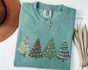 Könnte beinhalten: Ein türkisgrünes T-Shirt mit vier illustrierten Weihnachtsbäumen, die mit Lichterketten geschmückt sind. Die Bäume sind mit gelben, roten und weißen Lichtern geschmückt. Eine silberne Halskette mit einem kleinen Anhänger liegt auf dem T-Shirt.