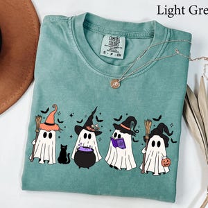 May include: Light green t-shirt with a graphic of four cartoon ghosts wearing witch hats and holding a broom, a cauldron, a book, and a pumpkin. A black cat is also in the graphic. The shirt has a Comfort Colors tag.
