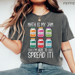 May include: A charcoal gray t-shirt featuring a graphic of six colorful jars labeled with math terms including equations, counting, numbers, addition, fractions, and problem solving, with the text MATH IS MY JAM I'M HERE TO SPREAD IT.