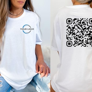May include: A white short-sleeved t-shirt featuring a blue and black circular logo on the front left chest with the text YOUR LOGO inside. The back of the t-shirt displays a large, square black and white QR code graphic centered.