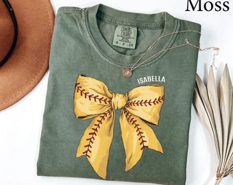 May include: Moss green t-shirt featuring a large yellow bow with red stitching, resembling a baseball. The shirt has the name "Isabella" printed in white. A silver necklace with a small pendant is draped across the shirt.