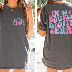 May include: Dark gray t-shirt with the text "DOUBLE DIGITS ERA" and a disco ball graphic on the front. The back of the shirt features the text "IN MY DOUBLE DIGITS ERA" with a smiley face, stars, and peace sign.