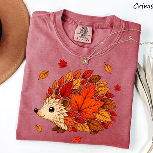 May include: A crimson short-sleeve t-shirt featuring a graphic of a hedgehog whose body is composed of various autumn-colored leaves in shades of orange, red, and brown. The shirt has a crew neckline and a Comfort Colors brand label at the neck.