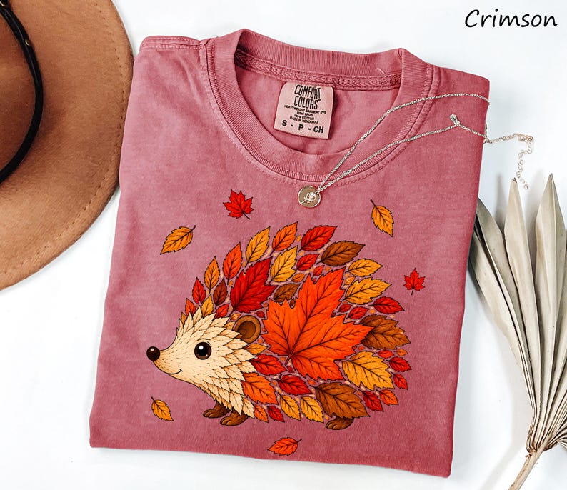 May include: A crimson short-sleeve t-shirt featuring a graphic of a hedgehog whose body is composed of various autumn-colored leaves in shades of orange, red, and brown. The shirt has a crew neckline and a Comfort Colors brand label at the neck.