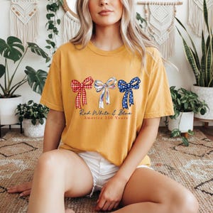 May include: A mustard yellow t-shirt featuring three bows in red gingham, white with blue stripes, and blue with white stars. Below the bows, the text reads Red White & Blue America 250 Years in a cursive script and a simple serif font.