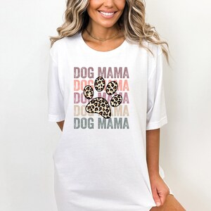 May include: White t-shirt with the text "DOG MAMA" repeated in a rainbow color scheme with a leopard print paw print in the center.