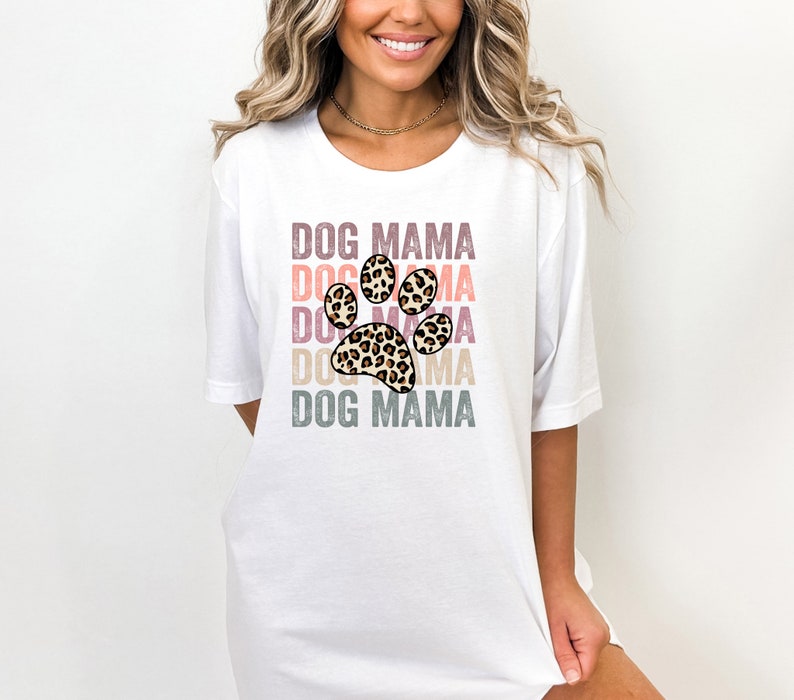 May include: White t-shirt with the text "DOG MAMA" repeated in a rainbow color scheme with a leopard print paw print in the center.