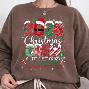 以下が含まれることがあります： 2026 Christmas Crew, A Little Bit Crazy, A Little Bit Loud, A Whole Lotta Loveという文字がプリントされた茶色のクルーネックスウェットシャツ。デザインには赤と緑の模様、サンタの帽子、キャンディケイン、小さなギフトが含まれています。
