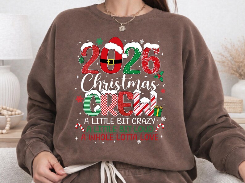 以下が含まれることがあります： 2026 Christmas Crew, A Little Bit Crazy, A Little Bit Loud, A Whole Lotta Loveという文字がプリントされた茶色のクルーネックスウェットシャツ。デザインには赤と緑の模様、サンタの帽子、キャンディケイン、小さなギフトが含まれています。