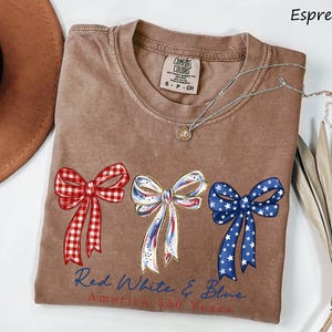 May include: An espresso-colored t-shirt featuring three decorative bows in red gingham, white with blue and red accents, and blue with white stars. Below the bows, the text reads Red White & Blue America 250 Years in blue and red script.