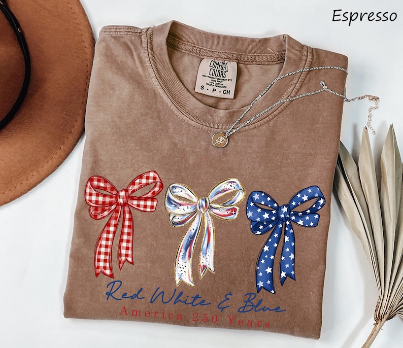 May include: An espresso-colored t-shirt featuring three decorative bows in red gingham, white with blue and red accents, and blue with white stars. Below the bows, the text reads Red White & Blue America 250 Years in blue and red script.