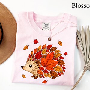 May include: A light pink t-shirt featuring a graphic of a hedgehog whose back is composed of various autumn-colored leaves in shades of red, orange, and brown. The shirt has a Comfort Colors brand label at the neck and the word Blossom is printed above.