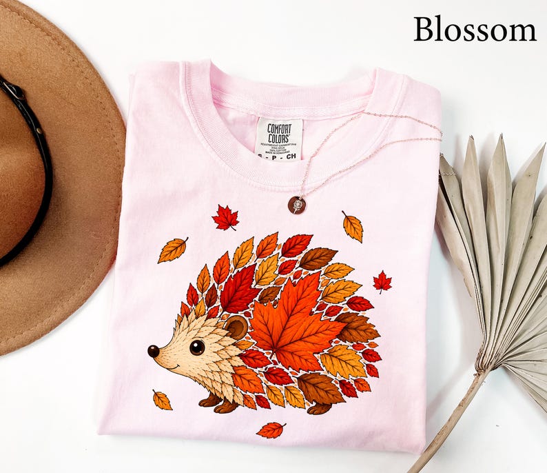 May include: A light pink t-shirt featuring a graphic of a hedgehog whose back is composed of various autumn-colored leaves in shades of red, orange, and brown. The shirt has a Comfort Colors brand label at the neck and the word Blossom is printed above.
