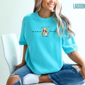 May include: A turquoise t-shirt with a graphic of Winnie the Pooh holding a honey pot, surrounded by bees. The text "mama bear" is above the image, with "EST. 2025" below. The word "LAGOON" is printed in the upper right corner.