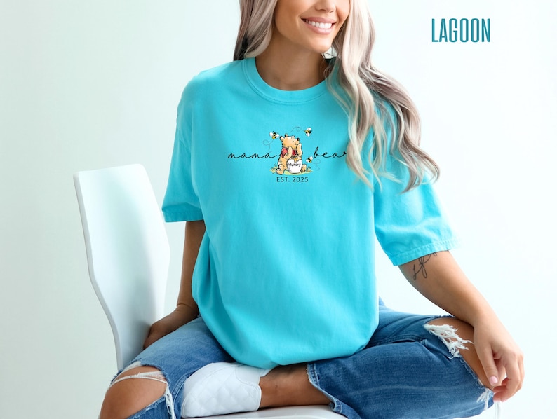 May include: A turquoise t-shirt with a graphic of Winnie the Pooh holding a honey pot, surrounded by bees. The text "mama bear" is above the image, with "EST. 2025" below. The word "LAGOON" is printed in the upper right corner.