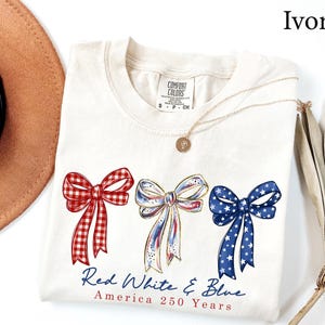 May include: An ivory t-shirt featuring three illustrated bows in red gingham, white with blue and red accents, and blue with white stars. Below the bows, the text reads Red White & Blue America 250 Years. The shirt has a Comfort Colors brand label.
