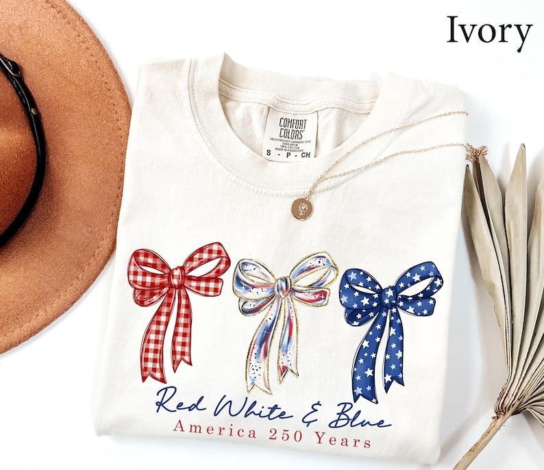 May include: An ivory t-shirt featuring three illustrated bows in red gingham, white with blue and red accents, and blue with white stars. Below the bows, the text reads Red White & Blue America 250 Years. The shirt has a Comfort Colors brand label.