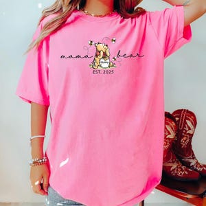 May include: Bright pink t-shirt featuring a Winnie the Pooh graphic with the words "mama bear" and "EST. 2025". The design includes Pooh hugging a honey pot with bees. The brand name "CRUNCHBERRY" is at the top left.