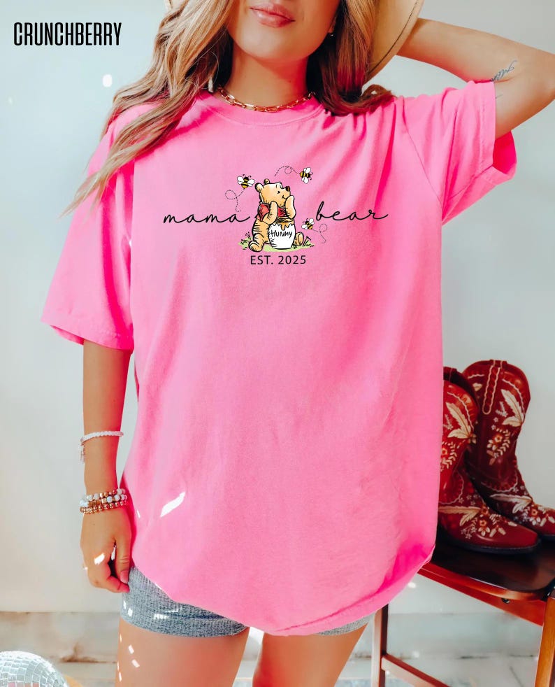 May include: Bright pink t-shirt featuring a Winnie the Pooh graphic with the words "mama bear" and "EST. 2025". The design includes Pooh hugging a honey pot with bees. The brand name "CRUNCHBERRY" is at the top left.