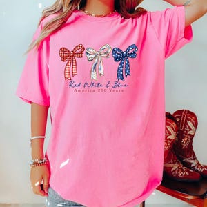 May include: A bright pink short-sleeve t-shirt featuring three bows in red plaid, white, and blue star patterns. Below the bows, the text reads Red White & Blue America 250 Years. The shirt has a crew neckline and a loose, relaxed fit design.
