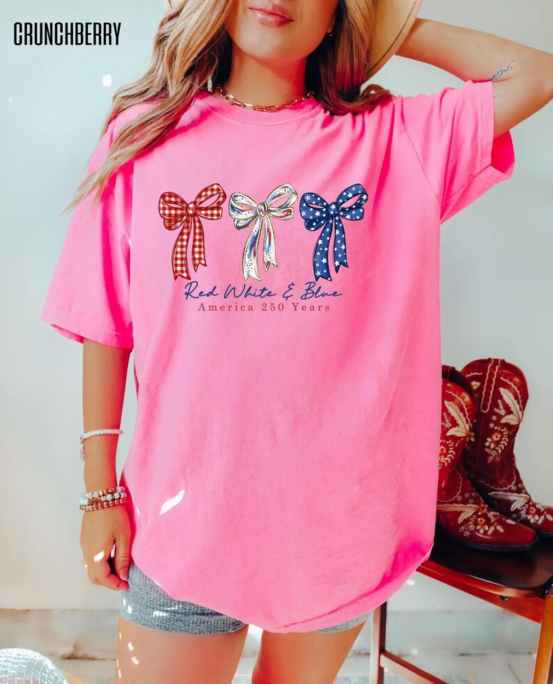 May include: A bright pink short-sleeve t-shirt featuring three bows in red plaid, white, and blue star patterns. Below the bows, the text reads Red White & Blue America 250 Years. The shirt has a crew neckline and a loose, relaxed fit design.