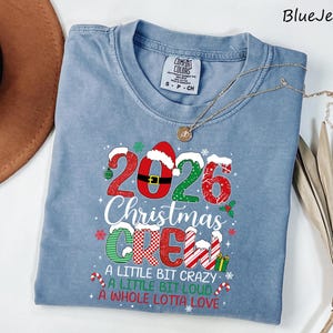 以下が含まれることがあります： 2026 Christmas Crew, A Little Bit Crazy, A Little Bit Loud, A Whole Lotta Loveという文字がプリントされた青いクルーネックTシャツ。デザインにはサンタの帽子、キャンディケイン、プレゼントなどのクリスマスらしい要素が含まれています。
