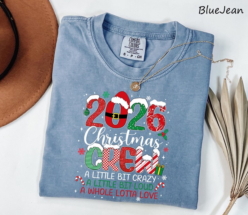 以下が含まれることがあります： 2026 Christmas Crew, A Little Bit Crazy, A Little Bit Loud, A Whole Lotta Loveという文字がプリントされた青いクルーネックTシャツ。デザインにはサンタの帽子、キャンディケイン、プレゼントなどのクリスマスらしい要素が含まれています。