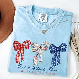 May include: A light blue t-shirt featuring three bows in red gingham, white with red and blue accents, and blue with white stars. Below the bows, the text reads Red White & Blue America 250 Years. The shirt has a Comfort Colors brand label.