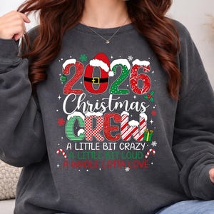 以下が含まれることがあります： 2026 CHRISTMAS CREW A LITTLE BIT CRAZY A LITTLE BIT LOUD A WHOLE LOTTA LOVEという文字がプリントされた、チャコールグレーのクルーネックトレーナー。赤と緑の模様、サンタの帽子、キャンディケインがデザインされています。
